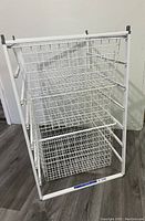Full view of the white metal wire basket storage unit showing all 3 baskets stacked vertically within the frame against a wall and floor background.