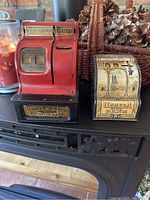 Front view of two early mechanical banks placed side by side on a black surface with pine cones in the background. The left is red Uncle Sam Register Bank with counter display, the right is USaveA Dime Bank with cream and blue decorative graphics.