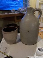 Wide view showing both stoneware crocks together on a white surface in indoor setting with TV and furniture in background.
