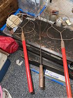 Three antique rug beaters laid on a wooden chest, showing red and wood handles with metal wire beater heads.
