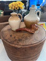 Two miniature stoneware jugs and one dog head pipe displayed on a round wooden surface with a flower pot and other household items in the background.
