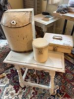 Large cream painted tin canister, small round canister, and rectangular spice box arranged on small white wooden table with woven rug and furniture background.