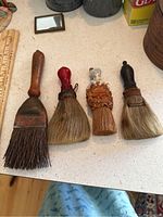 Four antique whisk brooms arranged in a row on a countertop, showing variation in handle materials and bristles.