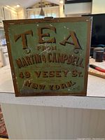 Front of vintage tea tin showing raised lettering 'TEA FROM MARTIN & CAMPBELL, 49 VESEY ST., NEW YORK' in gold and brown on green background.