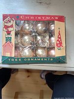 Full view of the box containing 12 round glass Christmas tree ornaments, visible pink and gold colors. Box is labeled Christmas Tree Ornaments.