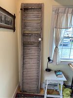 Full view of the tall narrow antique louvered door in a room corner showing the weathered gray wood and porcelain knob.