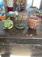 Front view showing all three flower frogs on a dark wooden surface: the green ceramic frog on the left, cast iron wire frog in the center, and cast iron basket-shaped frog on the right.