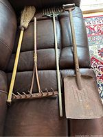 Four antique primitive child’s garden tools arranged on a brown leather couch showing full view of broom, wooden hay rake, green metal rake, and metal shovel.