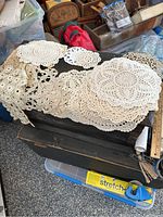 Top view of a pile of various hand crocheted doilies in cream color, showing different shapes and sizes with intricate patterns, placed on a dark surface