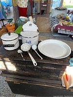 Six white enamelware pieces on a wooden chest including salt box, berry bucket, pie plate, and three miniature utensils