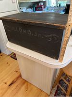 Photo of front side of the black painted wooden box showing the stencil 'H. LORD JR.' and part of the kitchen environment behind.