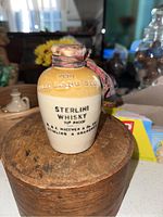 Miniature stoneware whisky jug with printed text on front showing brand and proof, original ribbon tied around the top, placed on a round wooden stand.
