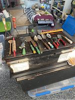 Overall view of the 18 vintage children's kitchen utensils laid out on a wooden chest, showing assortment of tools with wooden handles and metal parts in varied colors such as red, green, blue, white, and yellow.