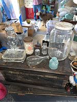 Photo showing eight vintage and antique glass bottles, jars, and a butterfly playing card box arranged on wooden furniture in a rustic room setting.