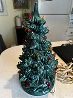 Front view of green Holland Mold ceramic Christmas tree with colorful small plastic bulbs on branches and plugged-in electrical cord for lighting.