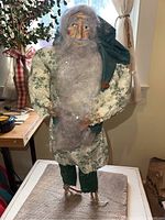 Front view of primitive homemade Santa doll standing on wood base, showing full figure with arms akimbo, clothing, and long gray beard.