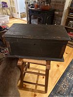 Front angled view of the primitive black painted wooden toolbox on a stool, showing overall box structure and lid.
