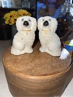 Pair of white mantlespaniel figurines on wooden surface, front view, showing textured fur and facial details.