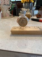 Front view of the small, round glass bottle with visible printed label and measurement ruler for scale.