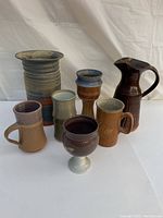 Seven assorted pottery pieces including vases, pitcher, mugs, and a goblet arranged on a white cloth background.