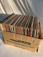 Cardboard box containing around 100 vintage LP vinyl records with spines visible showing varying colors and wear