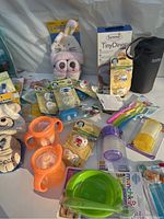 Photo showing assortment of baby feeding items including plates, bibs, cups, spoons, plush slippers, pacifiers and a bunny bonnet, mostly new with tags.