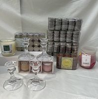 Photo showing two clear glass candlestick holders, many packs of silver tealight candles stacked, a pack of three mini jar candles from Bayside Candle Company, a white glass jar candle and a red jar candle.