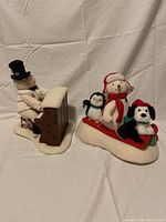 Snowman playing piano figurine on left and singing snowman with penguin and dog on right against white fabric background
