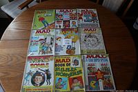 Top-down view of nine MAD magazines arranged on a round wooden table in three rows of three, showing colorful and illustrated covers.