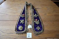 Full view of the antique Victorian tassel collar displayed flat on a wooden surface, showcasing purple velvet material, gold embroidery with floral and cross designs, and tassel edges.