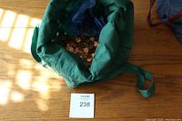 Green fabric bag filled with loose pennies on wooden floor with lot number card.