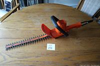 Full view of the Black & Decker electric hedge trimmer placed on a wooden table with the blade cover on and a lot number tag showing '210'.