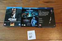 Front view of three Blu Ray box sets on wood floor: Die Hard Quadrilogy, Tron Two Movie Collection, and The Ultimate Bourne Collection with seller lot number visible