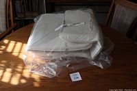 Folded off white/tan fabric replacement seat cover for IKEA POANG chair, sealed inside a clear plastic bag, placed on a wooden table.