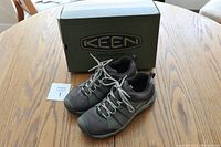 Pair of Keen women's hiking shoes in dark gray and black with laces on a wooden table next to a Keen branded shoebox.