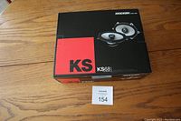 Closed box of Kicker KS68 speakers on wooden table with auction tag 154.