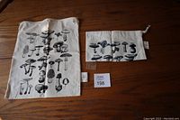 Two off-white fabric drawstring bags featuring black mushroom prints on a wooden surface, showing different mushroom designs and sizes.