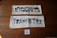 Two folded fabric tea towels lying flat on wooden surface. Top towel shows decorative black mushroom and foliage print. Bottom towel shows realistic mushroom illustrations with names in black.