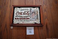Front view of the framed Coca Cola metal advertisement sign on a wood surface showing the detailed artwork and frame.