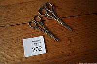 Two vintage grapevine scissors on wooden surface showing full view side by side with auction lot number card for scale.