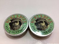 Two sealed tins of Burt's Bees Res-Q Ointment placed side by side on a white surface, showing front label with branding and illustration.