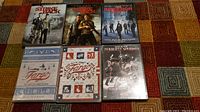Six DVDs displayed on a patterned carpet including Strike Back seasons 1 & 2, Inception, Fargo seasons 1 & 2, and Deadliest Warrior season 1.