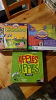 Photo showing three board games: Cadoo, Cranium bilingual edition, and Apples to Apples Junior, placed on a wooden table.