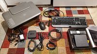 HP Deskjet 1055 printer, Apple TV 2nd gen, wired keyboard, wired mouse, Rogers TV box with remote and cables, all laid out on patterned carpet.