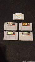 Five video game cartridges on a black surface showing one Nintendo 64 cartridge (F-1 World Grand Prix) and four Super Nintendo cartridges (PGA Tour 96, PGA Tour 97, Super Caesar's Casino, and NCAA Football).