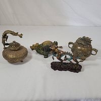 Four brass incense burners arranged side by side on a white surface, showing various shapes and designs including dragon and turtle.