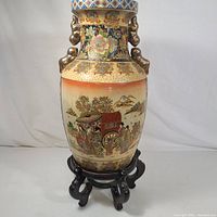 Full view of the jar on its wooden stand displaying the painted garden and geisha scene with applied handles.
