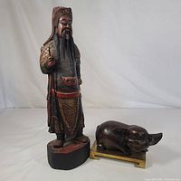 Two wooden figurines displayed on white background: an Asian warrior figure in detailed carved and painted wood, and a dark wooden pig on a brass base.