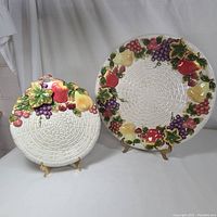 Two ceramic serving platters displayed on stands, showing their basket weave centers and fruit decorated rims.