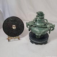 Photo showing the jade green stone incense burner with lid on a wooden pedestal next to the carved dark wooden circular decorative disk on a gold metal stand.
